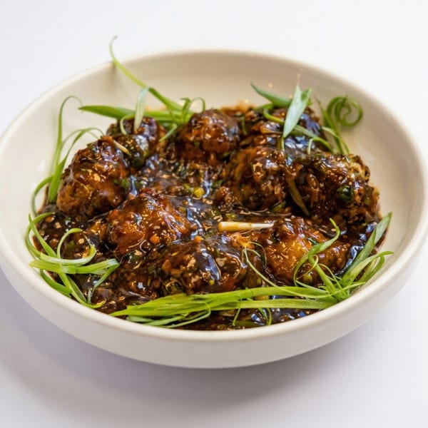 Close-up of Manchurian, dark brown sauce-covered vegetable balls garnished with green onions in a white bowl.